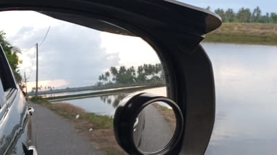 The image shows a view from inside a car, looking at the side mirror and the scenery outside. The car is on a paved road next to a body of water, likely a pond or rice paddy, given the flat landscape and rows of trees in the distance. The sky is overcast with hints of sunset. In the reflection of the side mirror, a road is visible with a telephone pole and some greenery. The overall scene suggests a rural or semi-rural setting. The location is identified as Pasi ie Leubeue, Indonesia.