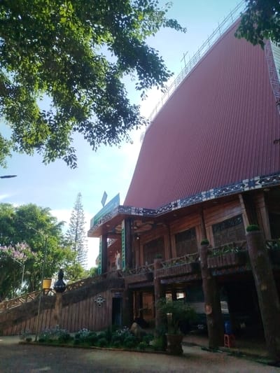 The image shows a building in An Phu Ward, Vietnam. The building has a tall, steeply pitched roof covered in red metal sheets. The lower section of the building features wood construction with ornate decorations. The roof is supported by scaffolding. A balcony is visible, and below it is a wooden-walled area with a dark doorway. There is a stairway with wooden railings leading up to the building. The surrounding environment includes trees, landscaping, and pavement.