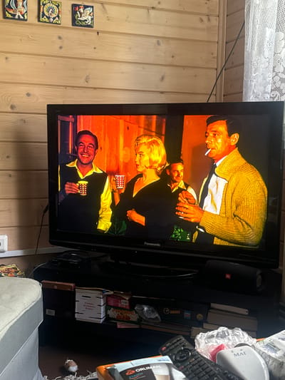 The image shows a television screen displaying a scene from a film or TV show. Three people are visible on screen. A man in the foreground, wearing a yellow sweater over a white shirt and dark tie, is laughing and holding a small checkered cup. Behind him and to the left, a woman with blonde hair, wearing a dark dress, is also holding a similar cup. To the far left, another man is seen wearing a patterned yellow cardigan and smoking a cigarette. The scene appears to be set indoors, possibly in a bar or a dimly lit room, with warm lighting.
To the left of the television, a wooden wall is visible, adorned with three small decorative tiles featuring mythological creatures or figures. To the right of the television, various objects are present on a shelf, including a black speaker with a red JBL logo, a white container with Russian text that appears to read "МАГ" (MAG), a remote control, a white plastic bag, and some boxes. One box has Russian text on it, which is partially legible and reads "РУССКОЕ АЛОЕ" (RUSSKOYE ALOYE) which translates to "Russian Aloe". Another white box with Russian text is also visible, with the text "В секрет. На вечерний. В буфет" (In secret. For the evening. To the buffet).
The overall context suggests a casual setting within a room, with the television as the focal point. The mood on screen appears lighthearted due to the man's laughter. The visible text indicates a Russian context for some of the objects in the scene. The lighting on the television suggests it might be evening or indoors with artificial lighting.