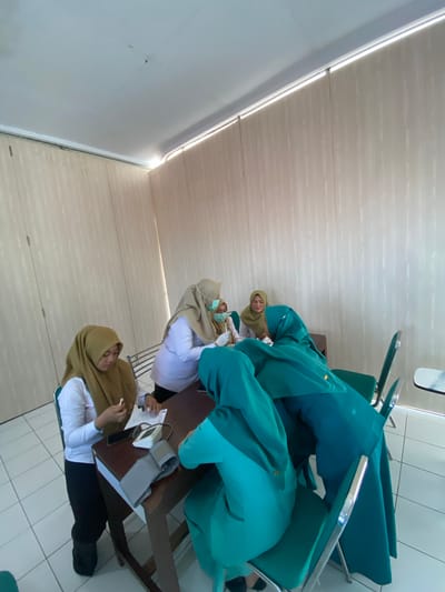In a room with cream-colored paneled walls and a white tiled floor, four individuals, all wearing headscarves, are gathered around a wooden table. One person, positioned behind the table and wearing a light purple top and a mask, appears to be administering a medical procedure. Another person, seated in front of the table and wearing a brown headscarf, holds a white object, possibly a pen or syringe, in their right hand and is looking down at a piece of paper. A blood pressure monitor with a cuff is visible on the table. Two other individuals, wearing teal headscarves and matching clothing, are seated in chairs to the right of the table. One of them is interacting with the person behind the table. The overall scene suggests a medical examination or a health screening taking place in an indoor setting, possibly a clinic or office in Margoyoso, Indonesia, given the context.
