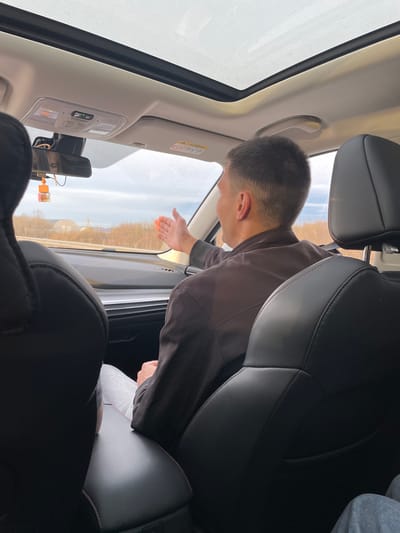 The image captures an interior view of a car, looking forward and to the passenger side, during what appears to be a clear, bright day. A male individual, seen from the back and side of his head, is prominently featured leaning out of the open passenger window, with his right arm and hand extended outward. He is wearing a dark brown jacket.

The car's interior features black leather seats, with subtle red stitching visible on the headrests. The ceiling and pillars are light beige or grey. An overhead console is visible, equipped with various buttons and indicator lights, one of which emits a green glow. A wide sunroof is partially visible, contributing to the bright lighting inside the car. A rear-view mirror is equipped with an attached dashcam or similar device, from which a small, orange rectangular air freshener or charm hangs. An "AIRBAG" warning label is present on the car's A-pillar near the windshield.

Beyond the car window, a bright sky stretches above a landscape populated by what appear to be bare trees lining a road or path, suggesting a rural or semi-rural environment, possibly during late autumn, winter, or early spring.

Parts of two other individuals are also visible: the torso of a person in the front passenger seat, wearing a light grey long-sleeved shirt, and the leg of someone in the back seat behind the leaning individual, wearing grey sweatpants. The scene depicts a passenger interacting with the outside environment from the vehicle. The location context is Kitsjanovo, Russia.