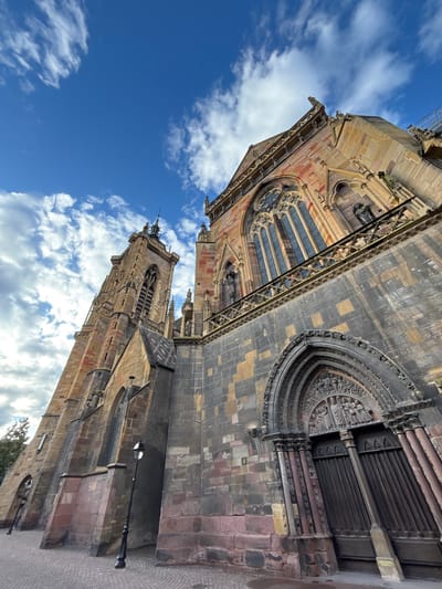 The photograph shows a low-angle, wide shot of the Collegiate Church of Saint Martin in Colmar, France. The sun is shining on a bright blue sky with scattered white clouds. The church's exterior is made of stone, with a mix of red and tan colors. The main facade features a large, arched wooden door with intricate carvings above it, flanked by ornate columns. To the left, a tall, narrow bell tower rises towards the sky, also constructed of stone. A street lamp stands on the cobblestone pavement in front of the church. The architecture is Gothic in style, with pointed arches and elaborate stone detailing visible on the windows and upper levels of the building.