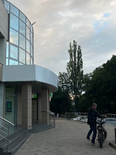 In the city of Belorechensky, Russia, on a cloudy day, a man walks his bicycle on a cobblestone sidewalk in front of a modern building with a curved glass facade. The building has large windows reflecting the sky and decorative lights along the top edge. A white awning extends over the entrance, supported by beige pillars. A sign is visible through a glass door. To the right of the building, a sidewalk slopes upwards towards a metal railing. Beyond the railing, parked cars are visible. Tall, slender trees and lush green foliage line the background. A second person, a woman in a pink outfit, walks in the distance on the sidewalk. The man is wearing a dark jacket, blue jeans, and dark sneakers. He is holding the handlebars of a black bicycle with a rack on the back.