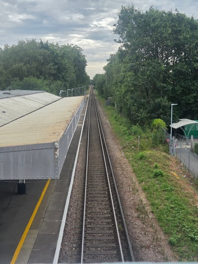 This is a high-angle, eye-level shot of a train track at Elmbridge station, United Kingdom. The sky is overcast with scattered clouds. On the left side of the image is a train station platform with a corrugated metal roof structure. The platform edge is marked with a yellow safety line. The train tracks run parallel to the platform and disappear into the distance. On the right side of the tracks, there is a grassy embankment bordered by trees. Further down the embankment, there is a covered bike rack area and a street light. The scene is quiet and appears to be during the day.