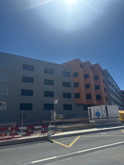 The image shows a modern building in Le Grand-Saconnex, Switzerland, under construction or renovation. The building features a grey facade with dark rectangular windows on the left, and an orange, stepped facade with similar windows to its right. Further to the right, a portion of another building with light-colored walls and numerous windows is visible. In the foreground, a street with yellow and white markings is present, bordered by a sidewalk and construction barriers. A white shipping container with "IMPLENIA" branding is positioned behind the barriers, along with construction signage and temporary fencing. The sky is clear blue with the sun prominently featured in the upper left corner, casting a bright glare.