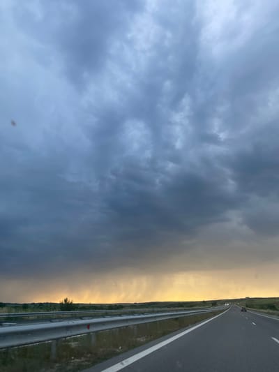 This image, presented vertically, should be viewed rotated 90 degrees clockwise to accurately depict the horizon. It captures a dramatic sky over a highway near Zlatna Livada, Bulgaria.
The upper portion of the corrected image is dominated by heavy, dark grey clouds, indicating stormy or overcast weather. These clouds display textured, undulating formations, particularly in the mid-left area. Towards the right side of the sky, a striking band of soft yellowish-orange light breaks through, suggesting either a sunrise or sunset, or a brief clearing in the turbulent weather.
Below the sky, on the right side of the frame, an asphalt highway stretches into the distance. It features clear white lane markings – a solid line defining the shoulder and dashed lines separating lanes. A metallic guardrail runs parallel to the road on its right side, beyond which lies a narrow grassy verge and a flat, open landscape dotted with sparse vegetation and a few small trees in the distance. Several indistinct vehicles are visible on the far stretch of the highway, marked by their lights. A small red reflector is attached to the guardrail. A slight, indistinct brownish smudge is noticeable in the lower-left sky area.
The scene conveys a sense of travel and the raw power of nature, with the contrasting light and dark skies creating a moody atmosphere. No discernible text is visible in the image.