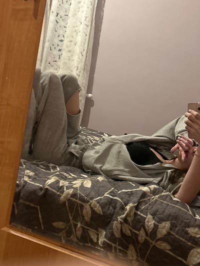 A person is reflected in a mirror. They are wearing a gray sweat suit with a black top underneath. The pants are ripped at the knee. The person is holding a phone. To the right of the person is a large, patterned pillow. The bed beneath the person has a white and light blue cover. The mirror's frame is made of light-colored wood. The wall in the background is a pale purple.