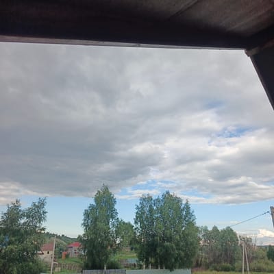 This image depicts a cloudy sky over a rural landscape in Podlubovo, Russia. The foreground is partially obscured by a dark, overhanging structure, possibly a roof or canopy, suggesting the photo was taken from indoors or a covered outdoor area.
The sky is filled with a mix of gray and white clouds, with patches of blue sky visible, indicating a partly cloudy or overcast day. The clouds are varied in texture, with some appearing dense and dark, while others are lighter and more scattered.
Below the sky, a line of trees, primarily deciduous with green foliage, stretches across the middle ground. Among the trees, some houses with red roofs are visible, along with fences and what appear to be utility poles and wires, suggesting a residential area within a rural setting. The vegetation is green and lush, typical of a warmer season. In the background, rolling hills are visible, covered with trees.
There is no discernible activity or event taking place; the scene appears to be a still landscape. The overall mood is somewhat somber due to the overcast sky, but the presence of green foliage and patches of blue sky offers a sense of natural beauty and calm. The time of day is likely daytime, given the lighting conditions, but the cloud cover makes it difficult to determine precisely. No text is visible in the image.