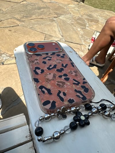 An outdoor scene in Olkaria ward, Kenya, depicts a mobile phone resting on a distressed white wooden surface, likely a bench or table, under bright sunlight. The phone is an iPhone, identifiable by its triple-lens camera array on a reddish-brown module with a metallic gold or silver trim. It is encased in a clear protective case featuring a salmon pink base with black and glittery rose gold leopard print spots. Attached to the bottom of the phone case is a lanyard-style phone charm composed of silver metallic beads (round and oval), black opaque round beads, clear translucent round beads, a silver metallic butterfly-shaped charm, and a black flower-shaped charm with a light-colored center.
To the immediate right, a tanned human leg, clad in a white two-strap sandal with a light-colored sole, is visible. This indicates a person is seated nearby. Partially visible behind the leg is an object with red and white stripes, consistent with a chair or bag. The ground surface is paved with irregular flagstones in natural earthy tones. The lighting suggests a clear day with strong shadows.