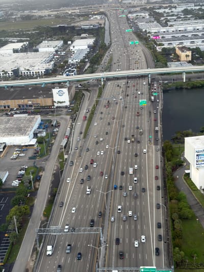 The image depicts a high-angle view of a multi-lane highway with heavy traffic, likely an interstate or major expressway. Numerous vehicles, mostly cars and trucks, are visible on the road. The highway is surrounded by urban development, including buildings, parking lots, and some green spaces. A body of water borders the highway on one side, while an elevated guideway or monorail structure runs parallel to it. Various road signs are visible above the highway lanes. Based on the architecture, landscape, and roadway design, the image likely captures a scene from Miami, Florida, United States.
