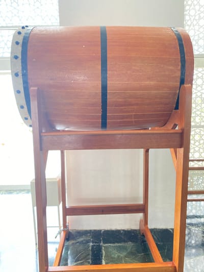 The image shows a large, wooden drum with black bands, supported by a wooden stand. The drum is cylindrical and appears to be made of light-colored wood planks joined together. There are black metal bands around the drum, and what looks like a skin stretched over one end, secured with dark-colored studs. The drum is elevated on a simple wooden frame, which has crossbars for stability. The background features a patterned screen or window with geometric designs, letting in soft light. The floor is tiled with dark and green marble. The overall scene suggests a cultural or ceremonial setting, possibly in a place of worship or a public building in Aluekambuk, Indonesia, given the context provided. There are no people or animals visible in the image, and no actions or interactions are depicted. The environment is indoors, illuminated by natural light.