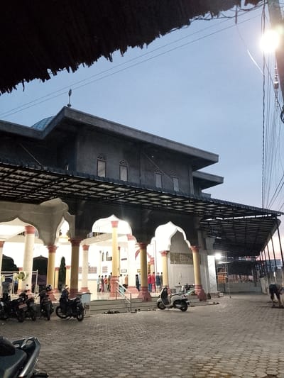 The image depicts the exterior of a mosque at dusk in Geulanggang Teungoh, Indonesia. A large, gray, two-story structure with a domed roof dominates the background. The mosque's entrance is flanked by white columns with red detailing, and a covered walkway extends from the building, supported by similar columns. Several motorcycles are parked in the paved courtyard in front of the mosque. A group of people, some dressed in traditional Islamic attire, are gathered near the entrance. The sky is a soft blue, indicating the end of daylight. The overall scene suggests a peaceful, community gathering at a place of worship.