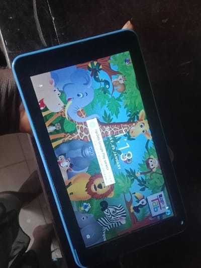 A tablet with a blue frame displays a children's cartoon-themed lock screen. The wallpaper features an elephant, giraffe, lion, zebra, monkey, and other animals in a jungle setting. A notification window prompts the user to update Google Play Services, indicating that certain apps won't function correctly without the update. The screen shows the time as 1:13 and the date as Sunday, January 25. A "Legacy Tab" icon is visible in the corner.