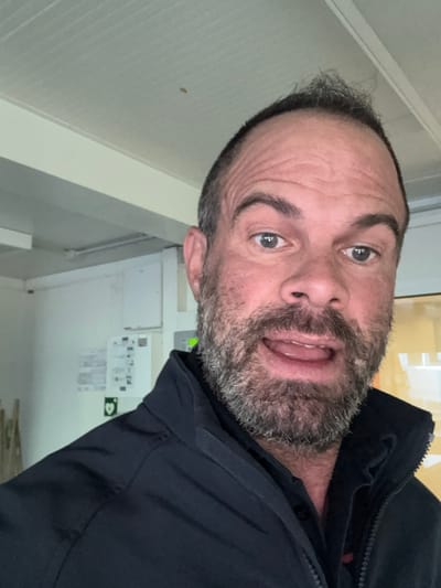 The image is a selfie of a middle-aged man with a receding hairline and a salt-and-pepper beard. He is wearing a black jacket. The background shows a white wall with what appear to be safety or informational signs. The location is potentially indoors, possibly an office or industrial space. The exact location in Diekirch cannot be determined with the available information.