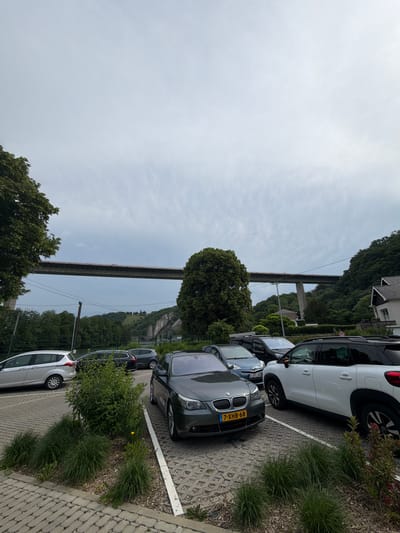 This image, apparently taken in Dinant, Belgium, features a row of parked cars along a brick and cobblestone parking area. The car closest to the viewer is a dark gray BMW, with its front visible. In the background, a bridge spans a valley or river, flanked by tree-covered hills. The sky is overcast.