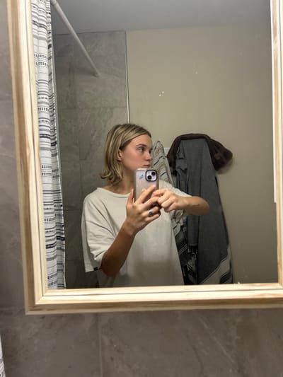 The image displays a mirror selfie of a young adult female in a domestic bathroom, located in Kingston, Canada. The subject, with shoulder-length blonde hair, is shown in profile, looking towards the right. She is wearing a loose-fitting, light beige short-sleeved top. She holds a light-colored smartphone vertically with both hands, positioned to capture her reflection; her left hand has a ring on the ring finger, and a small tattoo is visible on her outer left forearm. The smartphone's case is light-colored and features two small stickers, one resembling a butterfly. The mirror has a light wooden frame. The background reflected in the mirror includes grey rectangular wall tiles and a black and white striped fabric shower curtain on a silver tension rod to the left. To the right, a plain light beige wall is visible with a dark brown towel draped over a fixture and a grey and white striped towel hanging beneath it.