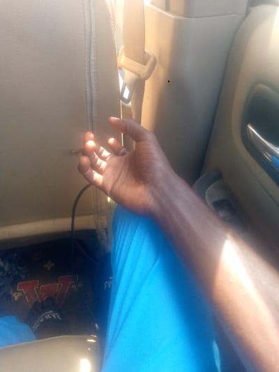 The scene depicts the interior of a vehicle, specified as being in Igede-Ekiti, Nigeria. A dark-skinned human's right arm, forearm, and open hand are prominently visible in the mid-frame, illuminated by direct sunlight. The individual's leg, clad in bright blue fabric, is visible below the arm. To the left of the hand, a beige car seat back is present, featuring a vertical seam and a small tear or scuff. Above and behind the hand, a beige seatbelt and its buckle are visible. The vehicle's interior paneling, also beige, is visible to the right, incorporating a chrome-colored door handle. On the floor, beneath the blue fabric, a dark object, possibly a bag, is partially visible, displaying white text that includes "BAHAMAS" and a red "W" logo on a patterned background. A black cable extends downward from the area near the car seat towards the floor.
