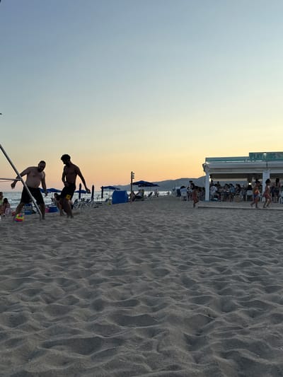 The image depicts a beach scene at dusk in Castelldefels, Spain, as indicated by the presence of the beach establishment named "PLAYEROS" which is a known venue in the city. In the foreground, a vast expanse of sand with rippled textures stretches across the frame, suggesting a recent tide or wind activity. Two men are engaged in a game of beach soccer in the middle ground. One man, with a lighter complexion, is dribbling a colorful soccer ball, while the other, with darker skin, appears to be attempting to defend or intercept. Both men are wearing dark shorts and are shirtless.

Behind them, the beach is dotted with beach umbrellas and lounge chairs, some of which are occupied. A building with a light-colored awning and the prominent sign "PLAYEROS" is visible on the right side of the image, housing several patrons seated at tables. Further in the distance, the sea is visible with gentle waves, and a faint silhouette of hills or mountains borders the horizon. The sky transitions from a clear light blue near the top to warm orange and yellow hues closer to the horizon, indicative of a sunset. Several other individuals, including children, are scattered across the beach, some walking and others sitting. The overall atmosphere is one of leisure and recreation.