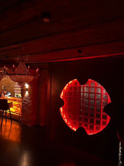 This is a dimly lit, high-angle interior shot of a room with exposed wooden beams on the ceiling and a brick wall in the background. The dominant color is red, cast by a large, red-lit window made of glass blocks in an unusual, scalloped shape, possibly resembling a bat or a stylized leaf. Below this window, a large, ornate crystal chandelier hangs, with multiple bulbs emitting a warm light. Further down, a person is partially visible, leaning over a counter or table, appearing to be working on something. The overall atmosphere is moody and somewhat mysterious, with warm lighting creating shadows and highlights. There are no visible signs of text or specific indications of the time of day or weather. The location is identified as Khamkhi, Russia, which might suggest a bar, restaurant, or a unique private dwelling.