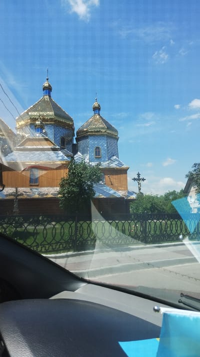 The image shows a view from a car interior, looking out through the windshield at an Orthodox church with blue and gold domes in Pavlivka, Ukraine. The church has a wooden facade and is surrounded by trees and a decorative black fence. A large cross stands to the right of the church. The sky is blue with some white clouds. The interior of the car is visible in the foreground, with the dashboard and a portion of the windshield reflected in the image.