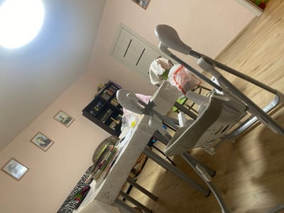 In a room with pale pink walls and a light wood floor, a brightly lit ceiling fixture casts a strong glow. A closed, light-colored door with dark horizontal lines is visible on the wall. In the foreground, a light gray baby swing with a white seat and tray dominates the scene. The swing is positioned at an angle, with its metal legs extending towards the floor. On a table covered with a white, lace-trimmed tablecloth, a stack of colorful books and what appears to be a zebra-patterned fabric are present. To the left of the swing, a dark wooden bookshelf is filled with books and various decorative items, including what looks like trophies. Three framed pictures are hung on the wall: two of individuals and one that is unclear. A plastic bag with red lettering is draped over the baby swing. The overall impression is a domestic setting, possibly a living room or dining area, with items suggesting the presence of a child. The location is identified as Афанасьево, Russia.