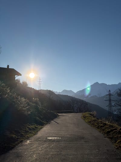 The image shows a road in Saint-André, France, with a clear blue sky partially obscured by the sun's glare. A building is partially visible in the foreground, silhouetted against the sun. Power lines and pylons stretch across the scene, traversing a mountainous landscape. The road itself is asphalt and extends into the distance. Vegetation lines the road's edge.