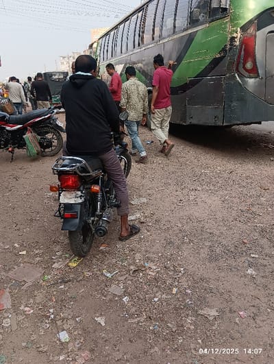 The image displays an outdoor street scene in Kaliganj, Bangladesh, dated 04/12/2025 at 14:07. The ground consists of unpaved dirt and gravel, heavily littered with paper, plastic, and other debris. In the foreground, a male individual is seated on a black motorcycle, facing away from the camera. He wears a dark long-sleeved top, dark trousers, and dark sandals, with a light-colored mask or collar around his neck. His right foot is on the ground. To the left, a second black motorcycle is parked, featuring a green and white striped bag suspended from its side.
In the midground, a large, dark green and black passenger bus, exhibiting noticeable dirt and wear, occupies the right half of the frame. Its rear section, taillights, and multiple side windows are visible. Approximately five to six adult males are present near and around the bus. One man, wearing a red shirt and light-colored pants, walks towards the bus with his right arm raised in a gesture. Another man in a patterned long-sleeved shirt and blue jeans is walking away from the bus. A third man in a red shirt stands alongside the bus. In the background, indistinct buildings are visible under a hazy sky, with overhead utility lines traversing the upper portion of the scene. The individuals' actions suggest routine street movement and interaction in proximity to public transport.