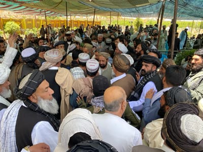 A large crowd of men wearing traditional Afghan clothing gather under a canopy made of white and green fabric. They are standing and talking to each other. Many of the men wear turbans or hats, and some have long white beards. The crowd is packed tightly together, and some men are holding their hands up in the air as they speak. The men are all of various ages, but the majority appear to be middle-aged or older.  Some men are wearing traditional Afghan vests and others are wearing long shirts or coats. 
This gathering is in the city of Zaker-E Sharif, Afghanistan, at a location with the coordinates (latitude: 31.547, longitude: 65.761). The scene is one of a large social gathering or a religious event.