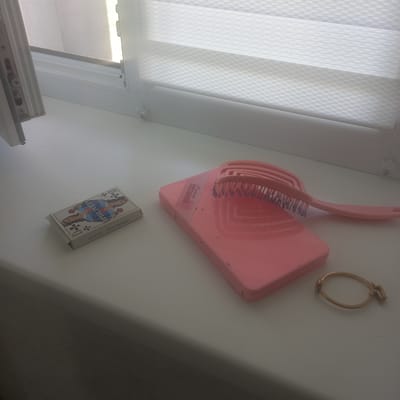 The image shows a windowsill in Cepeleuți, Moldova. On the windowsill, there are several objects: a deck of playing cards in a box, a pink dustpan with a pink brush resting on it, and a gold bracelet. The playing card box is decorated with an image of a queen. The dustpan and brush appear to be a set. The bracelet is delicate and appears to be made of gold. The windowsill is white and the background consists of a window with blinds that are partially open, allowing light to filter through. There are no people or animals visible in the image. The overall impression is one of domestic tidiness, with personal items neatly arranged.