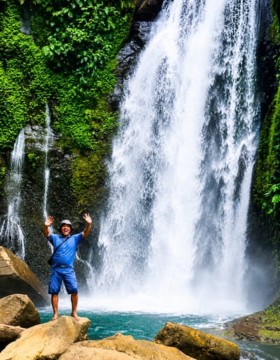 A man stands on a rock in front of a waterfall in Gunung Ambat, Indonesia. The man is wearing a blue shirt and blue shorts, and he has a hat on. He is smiling and has his hands up in the air. The waterfall is tall and wide, and it is surrounded by lush green vegetation. The water is clear and blue, and there are rocks in the foreground. The image appears to be taken in daylight.