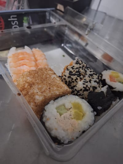 A plastic container holds a variety of sushi rolls and some cooked shrimp. The sushi includes several types: some are rolled with seaweed on the outside and topped with black and white sesame seeds, while others are inside-out rolls with rice on the exterior. One roll clearly shows salmon and cucumber. Another has a light yellow filling, possibly avocado or another type of seafood. A piece of sushi is heavily coated in toasted sesame seeds. To the left of the sushi, there are several pieces of cooked shrimp, their pinkish-orange bodies clearly visible. The sushi and shrimp are arranged within a clear plastic takeout container. The background is blurred, but appears to be an indoor setting, possibly a kitchen or dining area, with muted lighting. There are no people visible in the image. The scene is a simple close-up of food prepared for consumption. The visible text on a black label on the left side of the container reads "YOSH" upside down and "bsh" right side up. There is no indication of the time of day, weather, or specific emotions conveyed. The location context is Mińsk Mazowiecki, Poland.