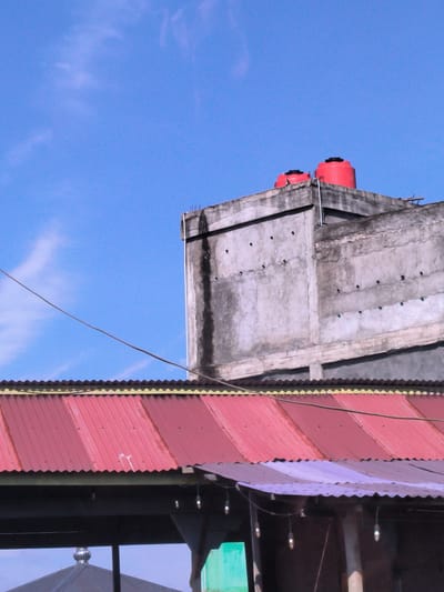 The image captures an upward view of various architectural elements against a clear, bright blue sky in Cotentung, Indonesia. Dominating the upper portion is a weathered, grey concrete building or structure, possibly unfinished, featuring multiple small, dark circular openings that might be vents or entries for birds. On its flat roof sit two prominent, round red water tanks. Metal rebar or rods protrude from the top edge of this concrete structure.
Below the concrete building, a large, corrugated metal roof, primarily red, stretches horizontally across the mid-ground. It shows signs of wear and discoloration. To the right and slightly below this, a smaller section of a purple corrugated roof is visible. From beneath the purple roof, several exposed light bulbs hang, suggesting an illuminated space below, possibly an outdoor stall or seating area.
A dark cable runs diagonally from the upper left to the middle right, crossing the blue sky above the red roof. A thin white pipe or conduit is visible vertically along the right side of the concrete structure. At the very bottom of the frame, dark structures are partially visible, including a dark, dome-shaped roof and a bright green panel or awning.
The scene depicts a sunny day with minimal clouds, indicated by the vibrant blue sky and strong light. There are no people visible, and no discernible text. The overall impression is one of utilitarian urban or semi-urban architecture.