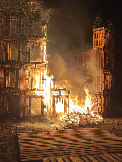 A towering structure composed of wooden pallets engulfed in flames against a dark night backdrop. The fire illuminates the stacked pallets, creating a vibrant orange and yellow glow amidst smoke. To the left, two evergreens appear to top off the construction. Sparks dance through the air, adding a dynamic effect to the intense inferno. Wooden planks are stacked adjacent to the main fire in the right corner.