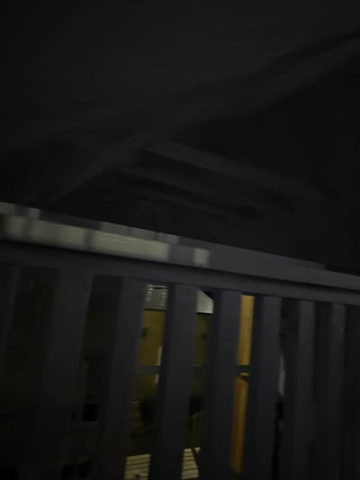 The image depicts a dimly lit, indoor scene. In the foreground, a light-colored railing runs horizontally across the frame, with vertical slats visible beneath it. Beyond the railing, a yellow object is partially visible, suggesting some sort of appliance or structure. The background is dark and indistinct, with diagonal lines suggesting a sloped ceiling or roof. There are no people or discernible actions. The location is identified as Doma 1096 Km, Russia.