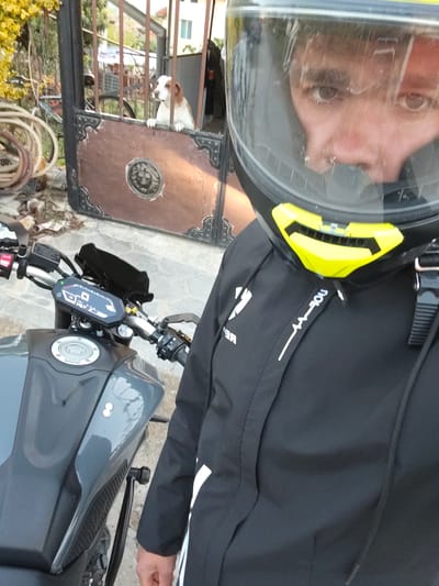 A person wearing a black motorcycle jacket and a black and yellow full-face helmet takes a selfie next to a gray motorcycle in Kulata, Bulgaria. The person's face is visible through the helmet's clear visor, showing their eyes and nose, with a neutral expression. The black jacket features white design elements, including what appears to be "YOU" written vertically on a strap near the neck, and possibly part of a brand name "REN..." on the left side.

The motorcycle is a dark gray color with visible handlebars, a digital instrument display showing "0" for speed, a "N" for neutral gear, and other indicators. Crash bars are visible on the side of the motorcycle.

In the background, a brown and white dog with short fur is peering over a dark brown metal gate, with its paws resting on the gate's top frame. The gate has an ornate circular emblem in the center. Behind the dog, coiled hoses and some yellow-leafed foliage are visible, suggesting an outdoor, possibly residential, setting. The lighting indicates it is daytime, with soft shadows. The overall scene captures a moment before or after a motorcycle ride, with the dog observing from behind the gate.