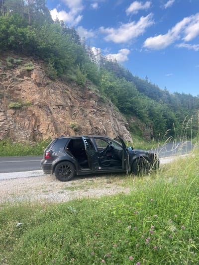 The image depicts a black Volkswagen Golf parked on the side of a road in a mountainous, forested area. The car's driver and passenger doors are open. The vehicle is situated on a gravel shoulder next to a paved road that curves into the distance. A steep, rocky cliff face with sparse vegetation rises to the left of the road. The area is covered in lush green grass and wildflowers, with tall trees and a power line tower visible on the hill in the background. The sky is blue with scattered white clouds. The location is identified as Pechinska, Bulgaria.