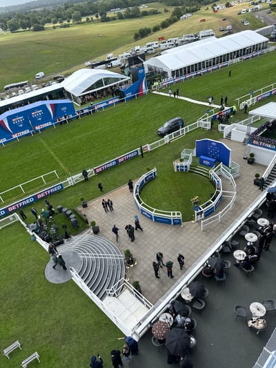 This high-angle, overhead image captures a large outdoor event at Epsom Downs Racecourse in Epsom and Ewell, United Kingdom, under an overcast sky. The scene is dominated by green grass and various temporary structures associated with the "Betfred Derby Festival."

In the foreground, a paved outdoor terrace features numerous dark-grey tables and chairs, mostly occupied by individuals or small groups. Many people are holding black umbrellas, some while seated. A white railing separates this terrace from the event grounds below.

The midground showcases the core event area. A circular grass enclosure, likely a winner's or paddock area, is bordered by white railings with "BETFRED THE DERBY FESTIVAL" branding. A central pedestal displaying flowers and a "WINNER" sign is visible within. Adjacent to this, a blue-paneled backdrop also branded "BETFRED DERBY" stands on a raised platform, with steps leading up to it. Various media equipment, including cameras on tripods and large equipment cases, are positioned on a paved section nearby. Several individuals, some in green jackets (possibly event staff or security) and others in suits, are present in this area. A dark-grey SUV is parked on a grassy patch behind a white fence line.

Stretching horizontally across the image is a section of the horse racing track, bordered by white fences and blue banners repeating "BETFRED." Beyond this, in the background, multiple large white marquees or tents are erected on green grass. The largest tent features numerous window panels along its length. Further back, on a gently sloping green hillside, a large collection of parked vehicles, including motorhomes, buses, and cars, suggests a spectator or support vehicle area. The distant background reveals expansive green hills dotted with trees under the grey sky.
