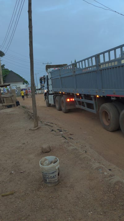 The image depicts a street scene, possibly in a commercial area. A large truck is parked along a dirt road. A utility pole with visible wires runs along the side of the road. A discarded NECOAT paint bucket sits on the ground near the truck. In the background, there are buildings, people, and various objects, suggesting a lively, populated area. The image suggests a roadside scene with evidence of human activity and commerce. No specific details confirm the location is in Masaka, Nigeria.