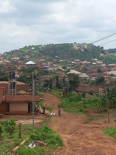 The image shows a dirt road winding up a hill in the town of Ita Oniyan, Nigeria, which is located at latitude 7.292 and longitude 5.167. The road is flanked by vegetation, including tall grass, bushes, and trees. There are numerous houses and buildings on both sides of the road, many of which are under construction. In the foreground, a man in a blue shirt walks on the road to the left, while two other men, who appear to be holding white plastic bags, walk on the right side of the road. In the background, a hill covered in foliage and several houses can be seen. The sky is overcast with clouds.