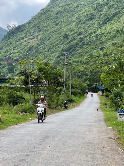 A person is riding a white scooter down a paved road. The road is flanked by lush green vegetation and a steep, heavily forested mountain on the left. The sky is overcast. The road curves to the right in the distance where several other motorcycles and a few people are visible. There are utility poles lining the left side of the road. A white sign with blue text, reading "KM 62 TRANG KIM KM 62," is visible on the right side of the road. The setting appears to be a rural mountainous area in Vietnam, consistent with locations like Cán Tỷ Commune.