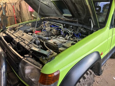 An overhead, slightly elevated, medium shot captures a bright green, off-road vehicle with its hood open in what appears to be a workshop. The vehicle's engine bay is exposed, revealing various components, wires, and a battery. The exterior of the vehicle, particularly the front fender and hood, has a textured, lime green paint finish. To the left, the vehicle's headlight is visible, as is a chrome-like pipe or fender. In the background, the workshop environment is suggested by wooden walls, tools, and overhead wiring. The scene is static and shows no people. The image is well-lit, with the primary light source appearing to be overhead, illuminating the engine compartment and the vehicle's exterior. The context of Lesnovo, Bulgaria is not directly deducible from the visual information within the image itself.