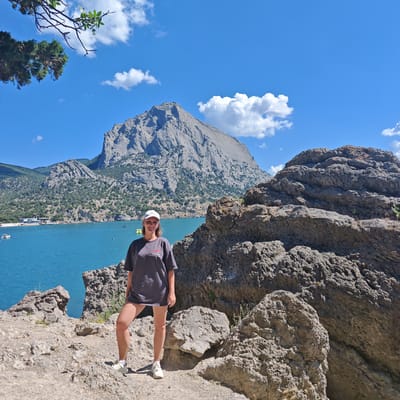 A young woman wearing a gray t-shirt, white shorts, and a white baseball cap stands on a rocky terrain, facing the camera. She is positioned in the foreground, with a turquoise body of water and a large, rocky mountain dominating the background. The water is calm, with a few boats visible in the distance. The mountain has a rugged, textured surface, and it is partially covered with greenery. The sky above is a clear blue with scattered white clouds. The overall scene suggests a scenic coastal location, likely a tourist destination, given the presence of boats and the natural beauty of the landscape. The location is identified as Novyi Svet, Ukraine, known for its stunning Crimean coast and mountains.
