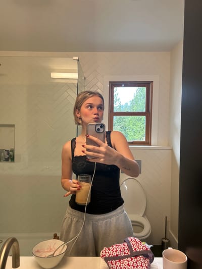 In a bathroom mirror selfie, a woman with short blonde hair is seen holding a beverage in a glass. She is wearing a black top with lace trim and grey sweatpants. Her nails are painted a dark red. The background includes white herringbone tile walls, a window showing greenery, a toilet, and various bathroom accessories. A glass shower door is visible on the left side of the frame.