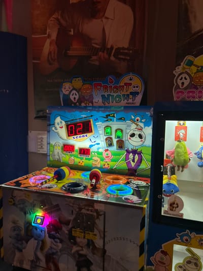 This image captures an indoor arcade scene, likely in Agios Dimitrios, Greece, featuring several entertainment machines and a wall poster.

The central subject is a vibrant arcade game named "Fright Night" by "FEILAI," which appears to be a whack-a-mole style game. Its colorful front panel depicts a Halloween or spooky theme, including a Jack Skellington-like character, ghosts, pumpkins, and a spider. Digital displays show "SCORE 120," "RECORD 113," and "TIME 000," suggesting the game has either just ended or is awaiting a new play. Two red-capped mallets are visible on the game's surface, positioned next to light-up holes. Above this game is a crane machine filled with various plush toys, including a green fuzzy creature and a blue and yellow shark-like character. Another sticker with "PBOO" is partially visible in the upper background.

To the left of the arcade game, a large poster is displayed, featuring a person playing an acoustic guitar. Visible Greek text on this poster reads "MIA TAINIA ADELPHON STEFAIO," which translates to "A film of brothers Stefanos," and then "TON STEFAN" and "TON KATER," likely referring to names.

The overall setting suggests an entertainment venue, possibly an arcade or family fun center. The lighting is artificial, common for indoor amusement establishments. The bright colors of the games contrast with the darker background, drawing attention to the activities.