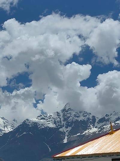 A majestic mountain range, capped with snow, dominates the background of this image. The sky above is a vibrant blue, interspersed with fluffy white clouds. In the foreground, the bright yellow, corrugated roof of a building angles across the bottom of the frame, with a hint of red trim and decorative white trim. The mountains are rugged and imposing, with dark, rocky slopes contrasting with the white snow. The overall scene suggests a remote, high-altitude location, likely within the city of Kardang, India, known for its dramatic Himalayan landscape. There are no people or active elements in the image, presenting a static, environmental view.