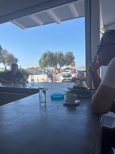 The image provides an interior view looking out onto Ornos, Greece, under a clear blue sky. In the immediate foreground, a dark grey counter surface extends across the lower portion of the frame. Arranged on this counter are a tall clear glass containing water, a circular light blue glass ashtray, a white ceramic coffee cup resting on a saucer, and a light-colored straw hat positioned on a blue patterned object. To the far right, a male subject, viewed in profile, wears dark sunglasses and holds a lit cigarette between his lips, with his right arm resting on the counter. To the left, a second male subject in a dark t-shirt is seated at an adjacent counter, with a small cup and a plastic water bottle visible near him. Beyond the interior space, the midground displays an outdoor area featuring light-colored paved surfaces, several green trees, and two parked vehicles—a white compact car and a red car. Several individuals are visible walking or standing in this area. The background showcases Ornos Bay, characterized by its blue water, a beach lined with numerous light-colored beach umbrellas and sunbeds, and several boats or yachts. A red and white structure stands prominently on the far side of the bay. The scene is brightly illuminated by natural daylight.