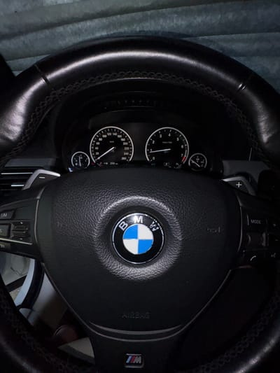 The image shows a close-up view of the interior of a black BMW, focusing on the steering wheel and dashboard. The steering wheel is made of leather and features the BMW logo in the center. Various buttons and controls are visible on the steering wheel, including paddle shifters, volume controls, and mode selection. The dashboard displays a speedometer and tachometer, with the speedometer showing approximately 259 km/h and the tachometer indicating just above 1000 RPM. The ambient light suggests it is daytime. The scene is likely captured inside a parked car, possibly in a residential area or garage, given the lack of any specific urban landscape visible. There is no information within the image to suggest a location in Novy, Russia.