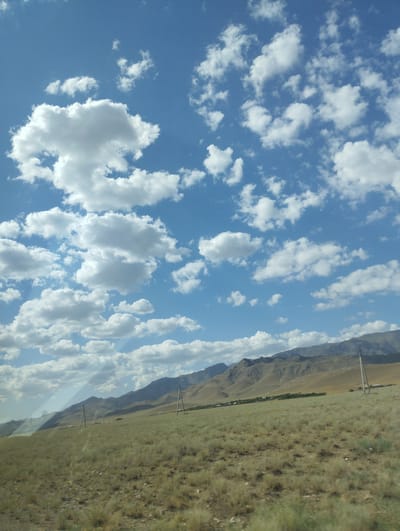 The image depicts a vast, dry, grassy plain stretching towards a range of low, barren mountains under a bright blue sky dotted with fluffy white clouds. The landscape is largely undeveloped, with a scattering of low-lying vegetation and a few utility poles visible in the distance. The terrain appears arid and somewhat desolate, suggesting a rural or remote location. There are no people or animals visible in the frame. The overall impression is one of a serene and expansive natural environment.