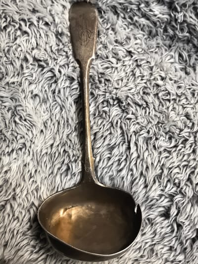 A close-up, overhead shot shows a silver ladle with ornate engraving on its handle, resting on a grey, fluffy, textured fabric surface. The ladle is positioned vertically, with the bowl at the bottom and the handle extending upwards. The fabric appears to be a blanket or throw, with a dense pile of wavy threads. The lighting is soft, creating subtle shadows on the fabric and highlighting the metallic sheen of the ladle. The scene appears to be an indoor setting, possibly a home. There is no visible connection to Ново-Талицы, Russia.