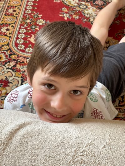 This is a close-up, vertically oriented photograph of a young boy lying down. He is looking towards the camera with a slight smile, revealing his teeth. His hair is brown and styled casually. He is wearing a white t-shirt with a repeating pattern of red text and circular green logos. The t-shirt has the visible text "SKIP PLAY FUN" printed on it.
The boy is positioned on a red, ornate carpet with intricate floral patterns in yellow, white, and black. Next to him, to his right, is a light-colored, textured surface that appears to be a blanket or rug. The background on the left shows a portion of the boy's arm and a dark gray t-shirt or garment he is wearing.
The lighting suggests it is daytime, possibly indoors given the presence of the carpet and blanket. The overall emotion conveyed is relaxed and cheerful. The location context provided is Радваніцкі сельскі Савет, Belarus.
